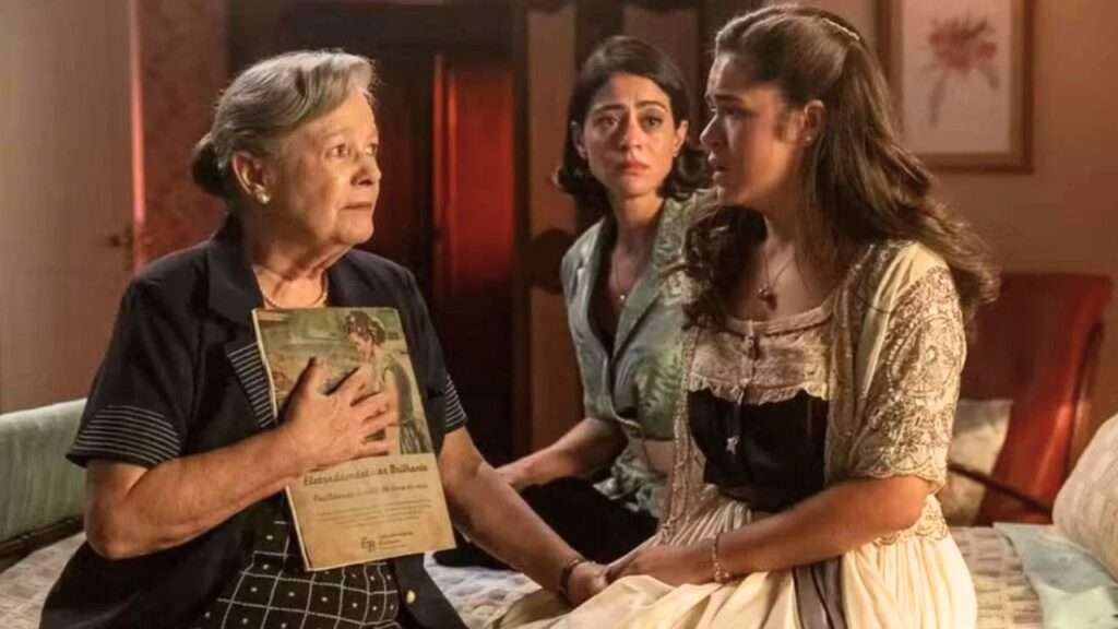 Arlete revela a Bia que é sua avó, segurando uma revista antiga como prova, enquanto Clarice observa emocionada a cena.